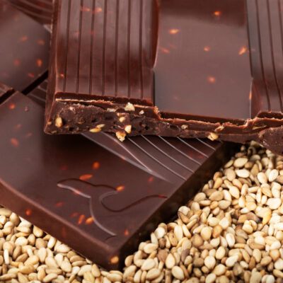 Tablette de chocolat noir 76 % & sésame soufflé de La Baleine à Cabosse dans son emballage, posée à côté d'un moule en forme de tablette rempli de grains de sésame