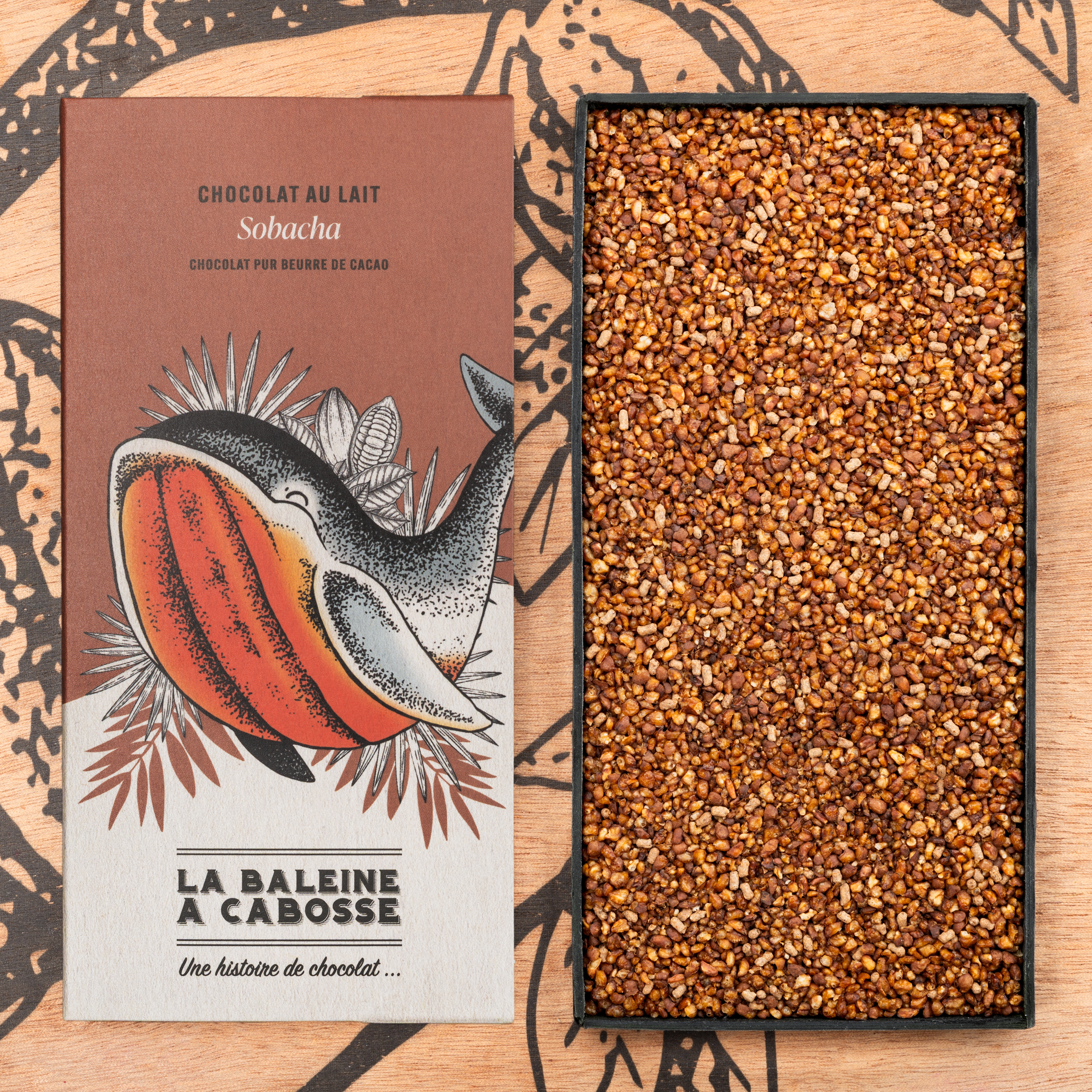 Tablette de chocolat au lait 50% de cacao & sarrasin grillé 1 Tablette de chocolat au lait 50% de cacao & sarrasin grillé de La Baleine à Cabosse dans son emballage, posée à côté d'un moule en forme de tablette rempli de sarrasin grillé