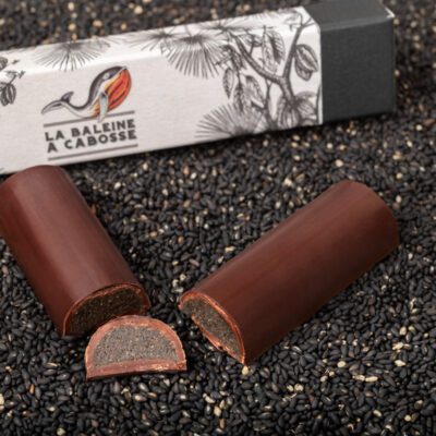 Chocolatier à Marseille - Chocolaterie artisanale La Baleine à cabosse 8 La barre fourrée au praliné de sésame noir coupée en deux et posée à côté de son emballage, sur une couche de sésame noir